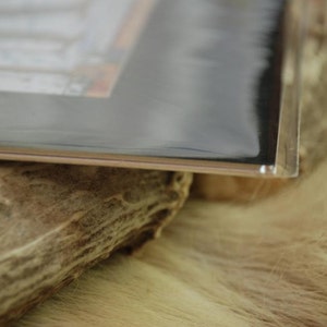 Puede incluir: Una funda de pl&aacute;stico transparente y rectangular con interior negro, apoyada sobre una superficie texturizada de color marr&oacute;n claro. La funda cubre parcialmente una superficie peluda de color claro y un trozo de madera.