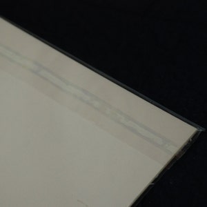 Puede incluir: Vista en esquina de un papel o tarjeta de color beige claro, sellado en una funda de pl&aacute;stico transparente. La funda de pl&aacute;stico tiene una cinta adhesiva fina y transparente a lo largo del borde. El fondo es negro.