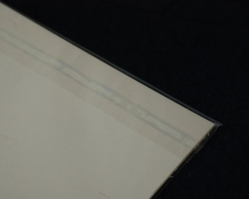 Puede incluir: Vista en esquina de un papel o tarjeta de color beige claro, sellado en una funda de pl&aacute;stico transparente. La funda de pl&aacute;stico tiene una cinta adhesiva fina y transparente a lo largo del borde. El fondo es negro.
