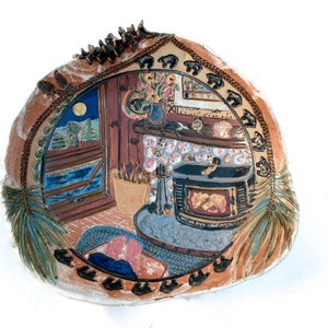 May include: A ceramic plate with a hand-painted scene of a cozy cabin interior. The scene includes a wood-burning stove, a window with a view of a moonlit landscape, and a person sitting on a rug. The plate is decorated with a border of stylized pine trees and bear paw prints.