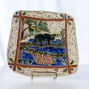 May include: A ceramic serving platter with a rustic design featuring a black bear and a raccoon on a log bridge. The platter has a cream background with brown, blue, and green accents.