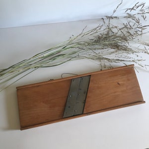 May include: A wooden vegetable slicer with a metal blade. The slicer is made of light brown wood and has a rectangular shape. The metal blade is silver and has a slightly curved shape.