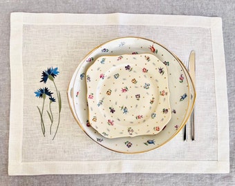 Puede incluir: Un mantel individual de lino blanco con un borde blanco y un diseño floral. El mantel individual tiene un juego de dos platos con un patrón floral y un borde dorado. Los platos están encima de un mantel individual de lino blanco.