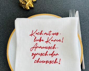 May include: A white linen napkin with red embroidered text, placed on a gold-rimmed plate with silverware. The text reads "Koch mit uns. liebe Karin! Armenisch. syrisch oder chinesisch!" A gold cherub figurine is in the background.