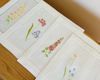 May include: Four white linen placemats with embroidered floral designs. Each placemat features a different flower in a different color: yellow, blue, pink, and white.