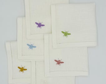 May include: A set of six square off-white linen napkins, each featuring a single embroidered bee in a different color: purple, lime green, light blue, mustard yellow, reddish-pink, and a subtle white bee on the bottom napkin.