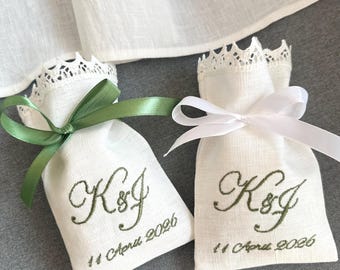 May include: Two small white linen bags with embroidered initials "K&J" and the date "11 April 2026" in green. One bag is tied with a green ribbon, the other with a white ribbon. Both bags have a lace trim.