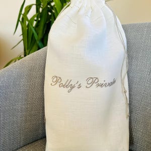 May include: A white linen drawstring bag with the embroidered text "Polly's Private" in a cursive font. The bag is sitting on a gray upholstered chair, with green foliage in the background.