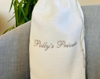 May include: A white linen drawstring bag with the embroidered text "Polly's Private" in a cursive font. The bag is sitting on a gray upholstered chair, with green foliage in the background.