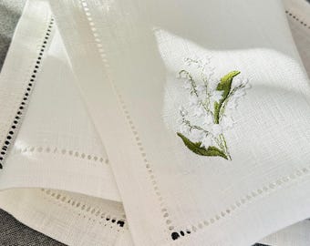 May include: A white linen napkin featuring an embroidered lily of the valley floral motif in green and white. The fabric has a visible woven texture and a decorative hemstitch border detail along the edges, creating a clean and structured appearance.