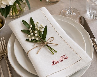 May include: A white linen napkin featuring a decorative hemstitch border and the embroidered red text Lia Ka. The napkin is folded and adorned with a small sprig of green leaves and white baby's breath flowers tied with a thin brown twine bow.