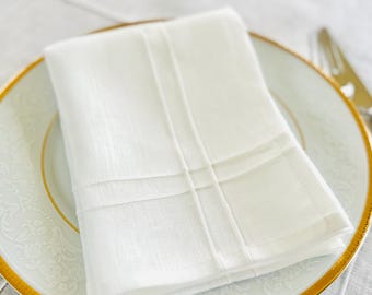 May include: A folded white linen napkin with vertical and horizontal stitched details rests on a white plate with a gold rim. The plate has a floral design. A fork is partially visible.