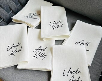 May include: A collection of white linen napkins, each with a different name in elegant black script. Names include "Aunt Abraham", "Uncle Phil", "Uncle Karl", "Aunt Kelly", "Aunt", and "Uncle Andy". The napkins are folded and arranged on a gray surface.