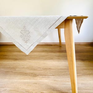 Puede incluir: Un mantel de lino beige claro con un diseño de árbol bordado. El mantel está sobre una mesa de madera con patas de color claro. El mantel tiene un borde cosido decorativo.