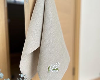 May include: A beige linen hand towel with an embroidered lily of the valley design. The towel is hanging from a door handle. The embroidery features white flowers and green leaves, adding a touch of nature to the home decor.