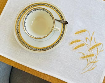May include: A white linen placemat with a wheat stalk embroidery design. A teacup, saucer, and spoon set with gold trim and a floral pattern are on the placemat. The table is made of wood.