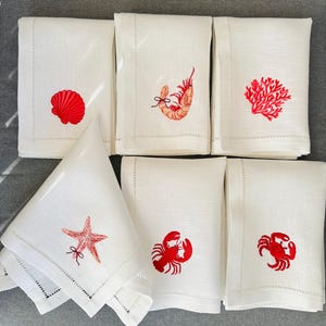 May include: White linen napkins with red embroidered sea life designs, including a seashell, shrimp, coral, starfish, crab, and lobster. The napkins are folded and arranged on a gray surface, showcasing a coastal theme.