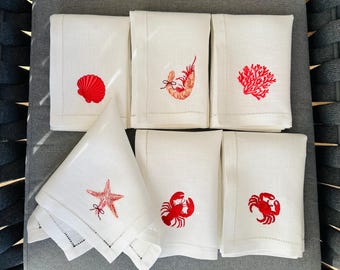 May include: White linen napkins with red embroidered sea life designs, including a seashell, shrimp, coral, starfish, crab, and lobster. The napkins are folded and arranged on a gray surface, showcasing a coastal theme.