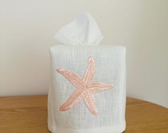 May include: A white tissue box cover with a light pink embroidered starfish design. The cover is made of a textured fabric and has a square shape. White tissues are visible at the top.