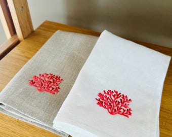 May include: Two folded linen towels, one light gray and one white, each embroidered with a coral reef design in bright red. The towels are neatly arranged on a wooden shelf, showcasing a coastal or nautical theme.