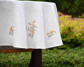 May include: White linen tablecloth with embroidered yellow flowers. The tablecloth is draped over a wooden table and the background is blurred greenery.