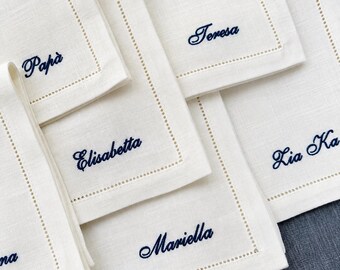 May include: A collection of off-white square linen napkins featuring a decorative hemstitch border. Each napkin is embroidered with a name in dark blue cursive script, including the names Papà, Teresa, Elisabetta, Mariella, and Lia Ka.