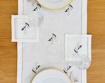May include: A table setting featuring a white linen table runner with embroidered lily of the valley sprigs. Two white plates with gold rims, silverware, clear glasses, and matching napkins complete the elegant dining arrangement.