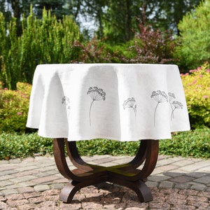 May include: White linen round tablecloth with a delicate floral pattern of black dill sprigs. The tablecloth is draped over a wooden table with a round top and four legs.