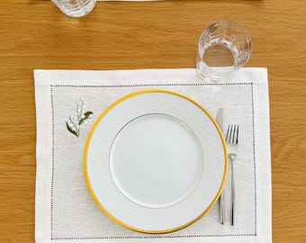 May include: Two white rectangular linen placemats featuring a delicate embroidered lily of the valley motif. Each setting includes a white porcelain plate with a gold rim and embossed pattern, a silver fork and knife, and a clear glass tumbler.