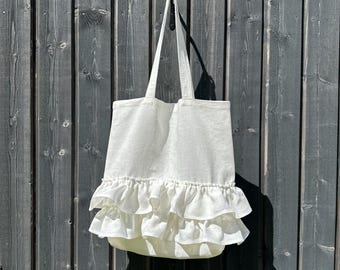 May include: A white tote bag featuring two horizontal layers of ruffled fabric trim along the lower section. The bag includes two long shoulder straps attached to the top opening, constructed from a plain, light-colored woven textile material.