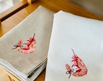 May include: Two linen napkins, one beige and one white, each embroidered with a red shrimp design. The shrimp has detailed shading and a black bow. The napkins are folded and rest on a wooden surface.