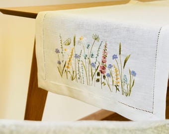 May include: A white linen table runner with an embroidered floral design. The embroidery features a variety of wildflowers in shades of yellow, pink, blue, and green. The runner is placed on a wooden table.