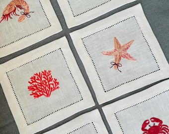 May include: Set of six white linen coasters with embroidered sea life designs. Each coaster features a different red embroidered design: a lobster, shrimp, starfish, coral, shell, and crab. The coasters have a dotted border.