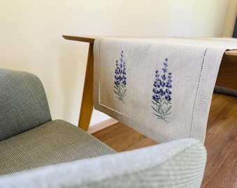 May include: A linen table runner with embroidered lavender sprigs. The runner is a natural beige color with two embroidered lavender sprigs in shades of purple and green. The runner is placed on a wooden table.