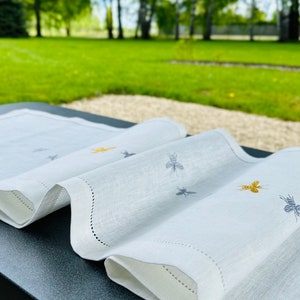 Puede incluir: Camino de mesa de lino blanco con diseños de abejas bordadas en amarillo y gris. El camino de mesa tiene un borde festoneado y está drapeado sobre una superficie oscura. El fondo presenta un césped verde y árboles.