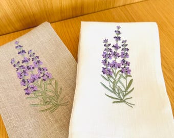May include: Two linen tea towels with embroidered lavender flowers. One towel is beige and the other is white.