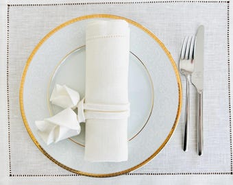 May include: Elegant table setting featuring a white plate with a gold rim, a rolled white napkin, and a folded napkin. Silverware, including a fork and knife, is placed to the right. The setting rests on a white placemat with a stitched border.