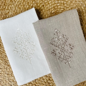 May include: Two folded linen napkins, one white and one taupe, each embroidered with a symmetrical, stylized tree design. The napkins are displayed on a woven, natural-toned surface.