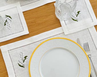 May include: A set of white linen placemats and napkins featuring delicate green and white lily of the valley embroidery, paired with white dinner plates with gold rims, a clear glass tumbler, a silver fork, and a silver knife.