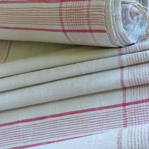 Pode incluir: Tecido branco sujo dobrado com um padrão xadrez vermelho. O tecido está parcialmente enrolado, revelando a borda e uma ligação de barbante natural. O design xadrez apresenta listras vermelhas horizontais e verticais.