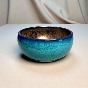 Peut inclure: Un bol en céramique rond présentant un extérieur dégradé turquoise et bleu sarcelle avec une texture mouchetée. L'intérieur du bol possède une finition métallique aux tons bronze avec un aspect marbré, contrastant avec la surface extérieure bleue.