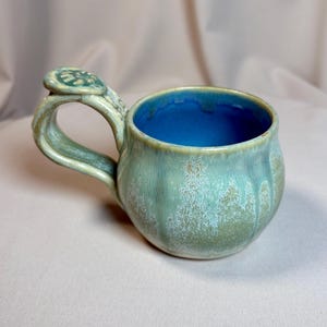 May include: A handcrafted ceramic mug with a distinctive design. The mug has a rounded form and a handle with a decorative feature. The exterior is glazed in green and blue, with a bright blue interior. Ideal for tea or coffee.