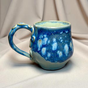 May include: A handcrafted ceramic mug with a blue and white glaze. The mug features a rounded form and a decorative handle. The interior is a pale green hue. Suitable for tea, coffee, or other drinks.