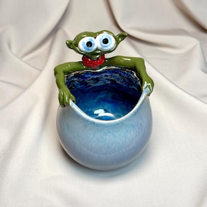 May include: A quirky ceramic bowl shaped like an alien, with a green body, large blue eyes, and a red neckerchief. The bowl is light blue with a deep blue interior, suitable for small items or as a decorative item.