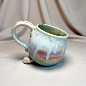 May include: A handcrafted ceramic mug with a distinctive design. The mug has a light blue handle with a textured pattern and a colourful glaze with green, red, and white hues. It rests on a small stone.