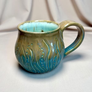Peut inclure: Mug en céramique fait à la main avec un design unique. L'intérieur du mug est turquoise et l'extérieur texturé présente un mélange de couleurs beige et turquoise. La anse est de couleur beige assortie. Le mug a une capacité d'environ 350 ml.