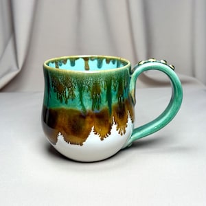 May include: A ceramic mug with a rounded body featuring a glaze that transitions from a bright turquoise interior to a deep forest green and brown drip pattern over a white base. The handle is green and features small, raised decorative bumps.