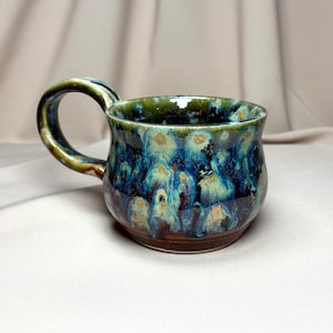May include: A ceramic mug with a rounded body and a curved handle, featuring a reactive glaze finish in shades of deep blue, dark brown, and light green. The surface displays a mottled, dripped pattern with lighter circular accents throughout.