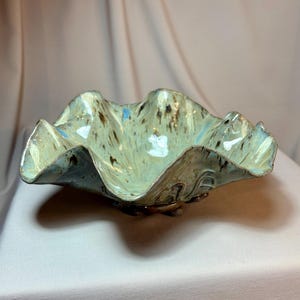 May include: A handmade ceramic bowl with a ruffled edge. The bowl has a light green base colour with brown and blue speckles. The rim is a darker blue. The bowl is a decorative piece.