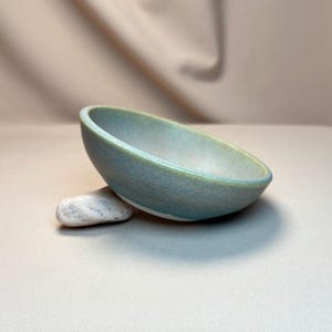 Peut inclure: Un bol en céramique fait à la main avec un glaçage bleu clair et vert. Le bol a une forme lisse et arrondie et est soutenu par une petite pierre blanche. Le bol est idéal pour servir des aliments ou comme pièce décorative.